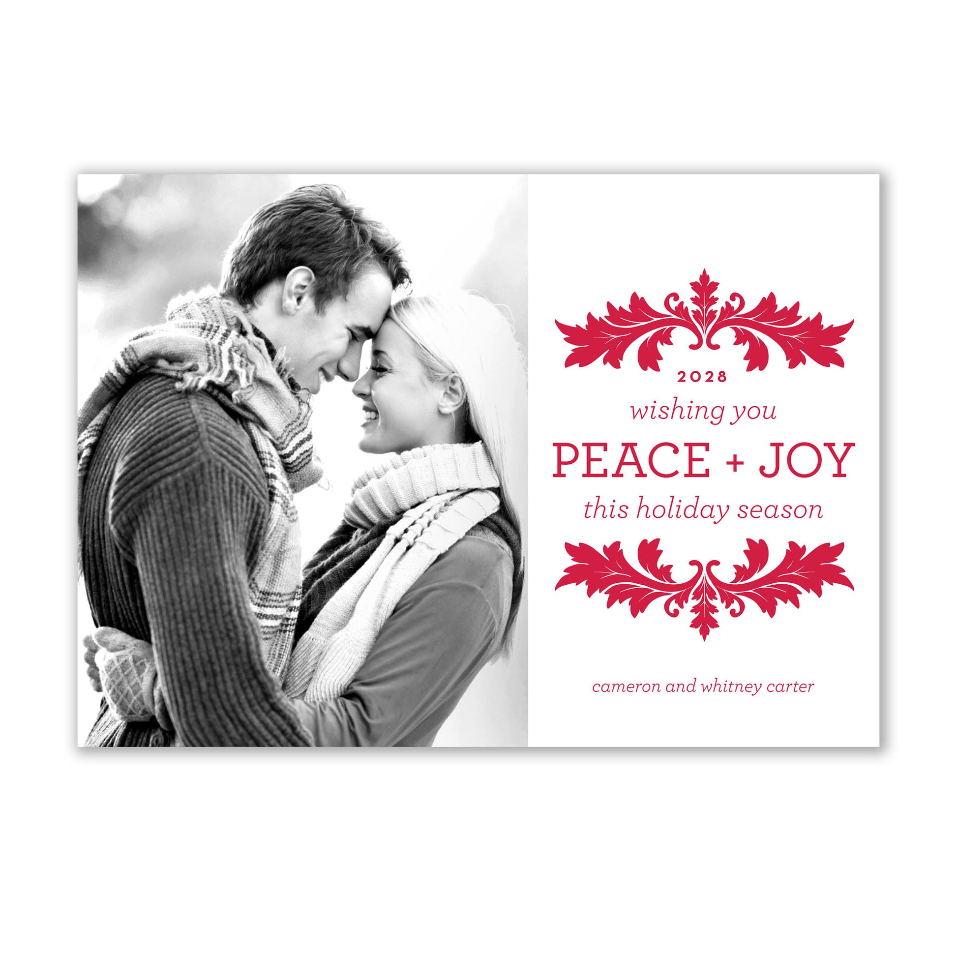 A couple in winter wear hugs outside next to the Noteworthy Holiday Bliss Photo Card, displaying a red holly design and the message, “2028 wishing you PEACE + JOY this holiday season cameron and whitney carter.”.
