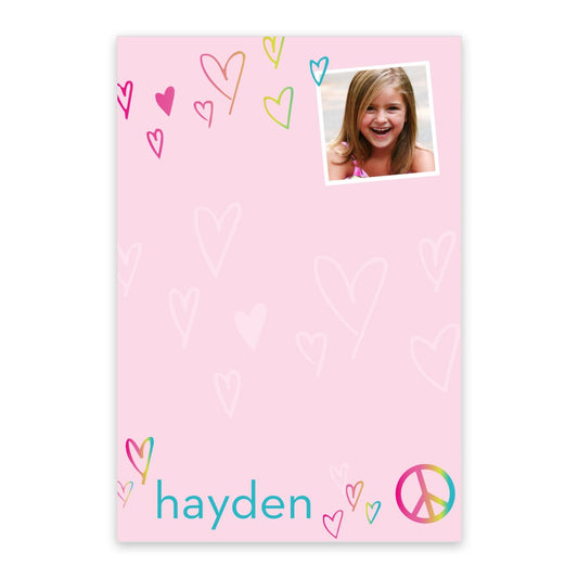 The Noteworthy Peace and Love Photo Personalized Notepad is pink, features doodled hearts, a small photo in the top right, and the name "hayden" with a peace sign at the bottom.
